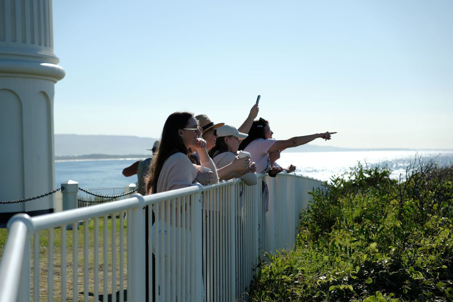 Group of women pointing at ocean views from a white railing near a coastal lighthouse.