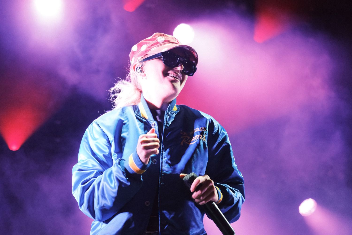 A performer in sunglasses and a cap sings on a smoky, colorful stage under red and purple lights.
