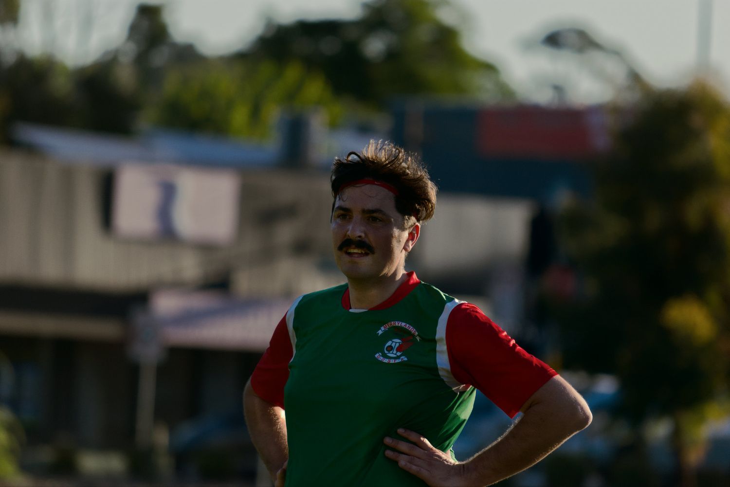 A mustachioed man in a green and red soccer jersey stands confidently on a sports field.