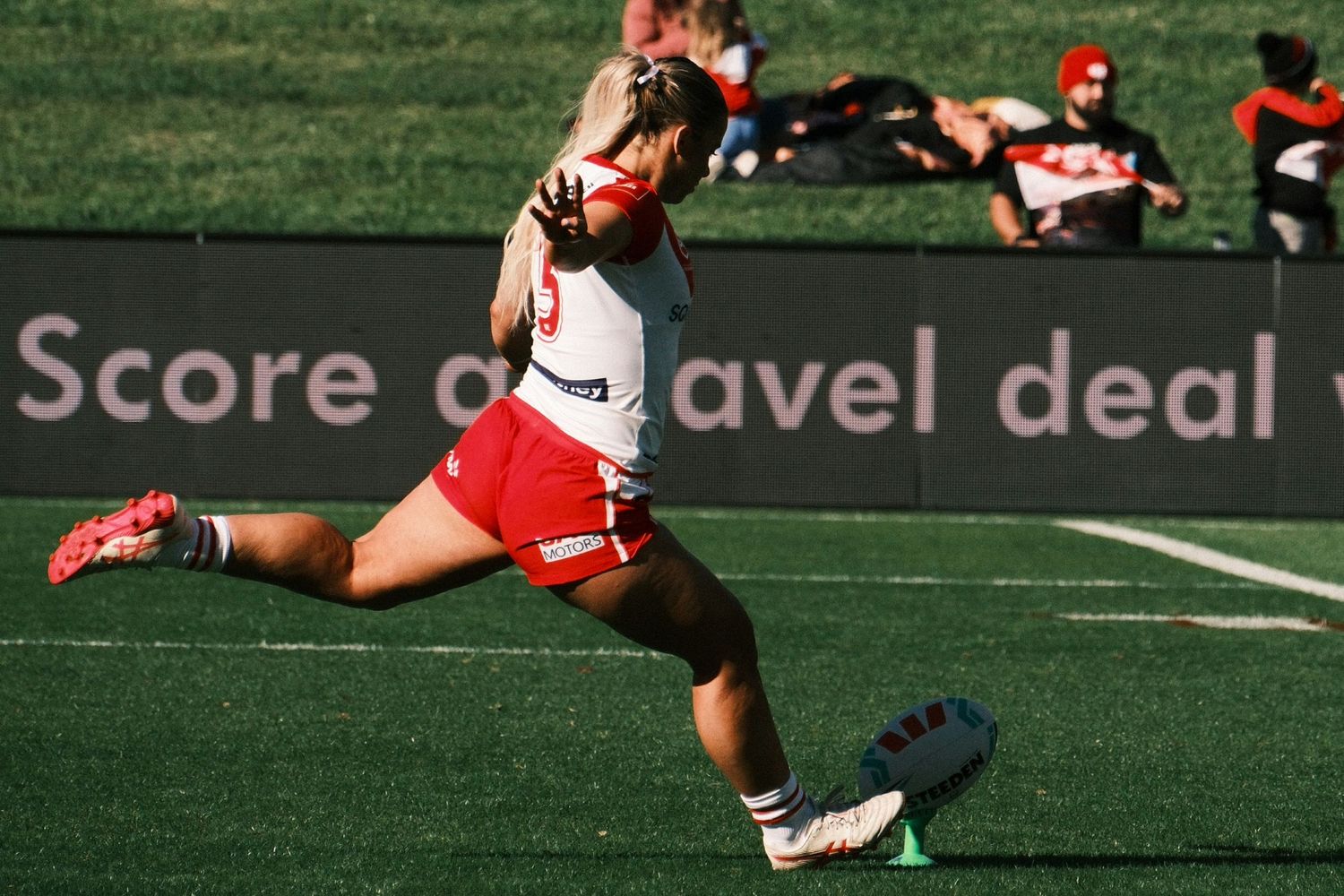 Female rugby player in red and white kit kicking the ball during a match on a green field.
