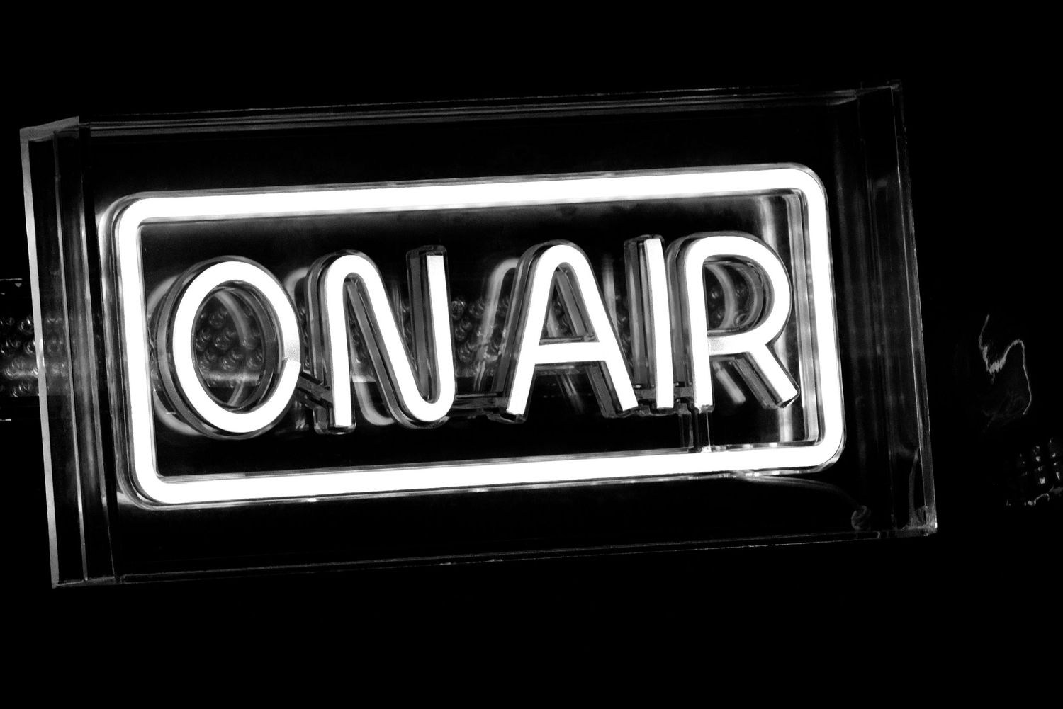 A glowing white neon ON AIR sign against a black background used in radio or recording studios.