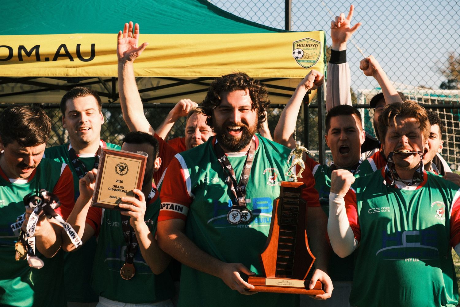 Soccer team celebrating championship win, holding trophy and medals in green jerseys outdoors.
