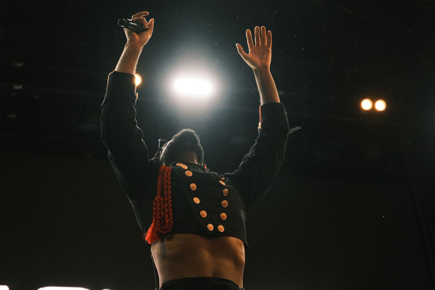 Performer on stage with arms raised, wearing a beaded jacket, backlit by bright concert lights.