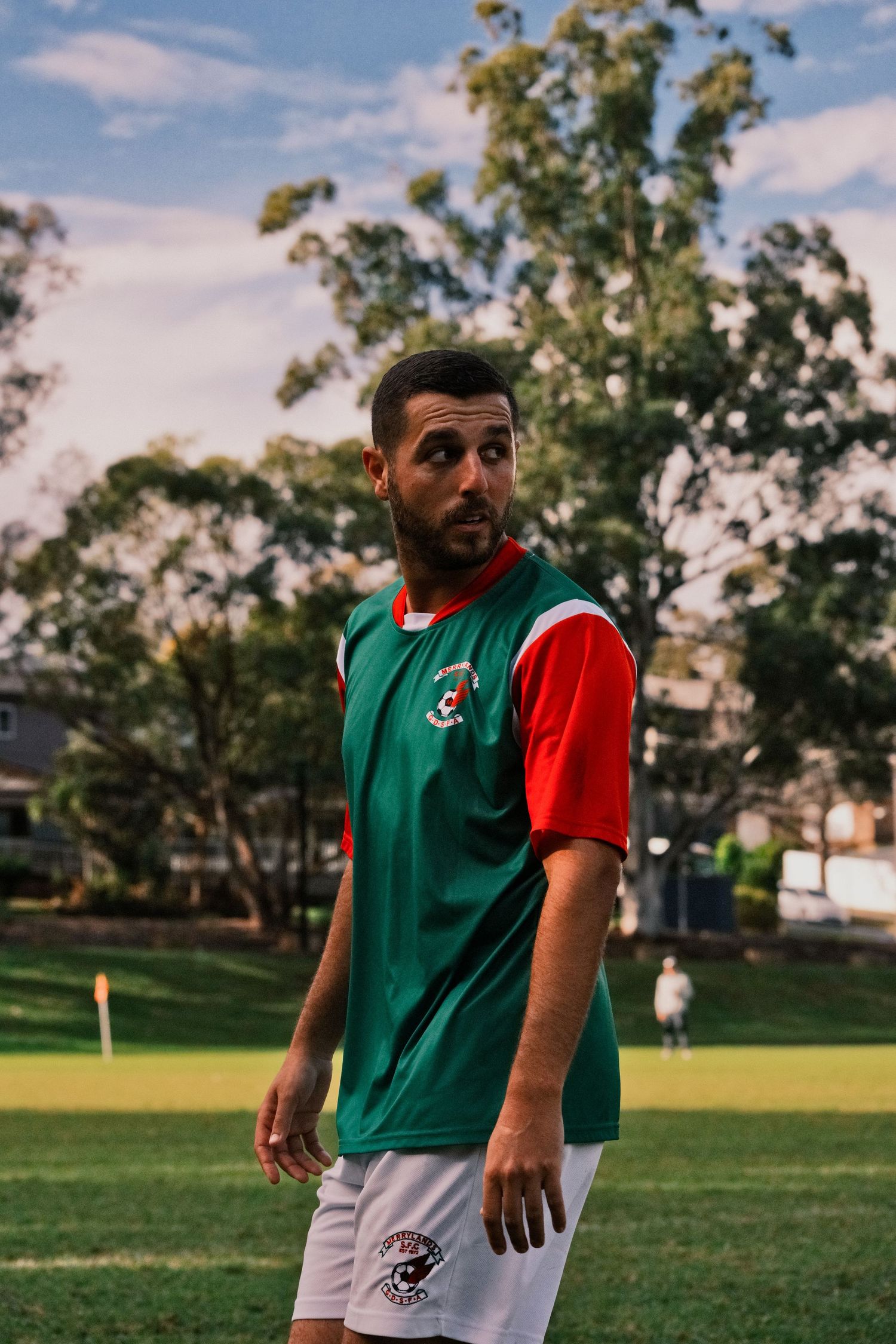 Male soccer player in green and red jersey stands on grass field with trees in background.