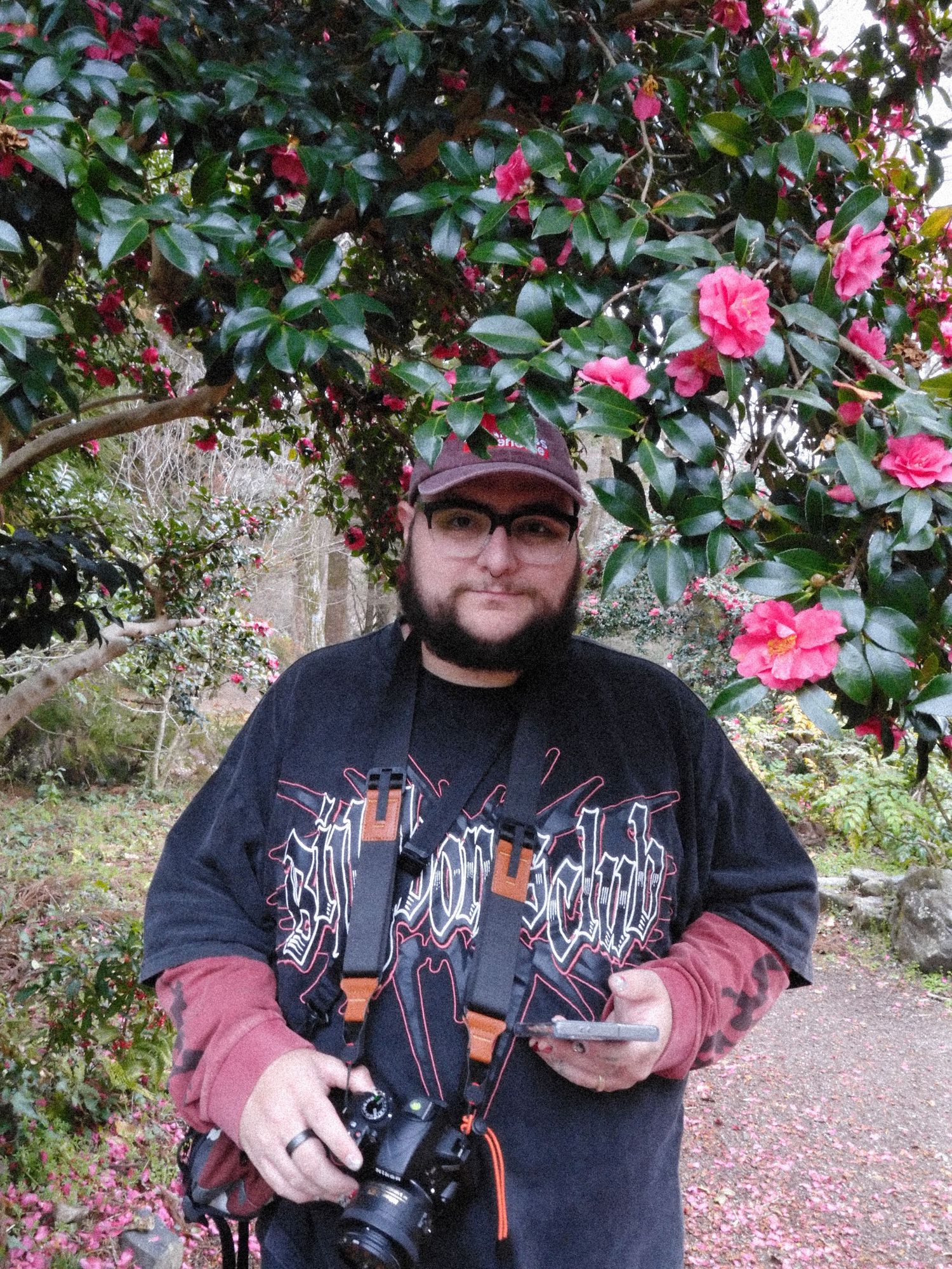Bearded photographer holding camera standing before blooming pink camellia flowers outdoors.