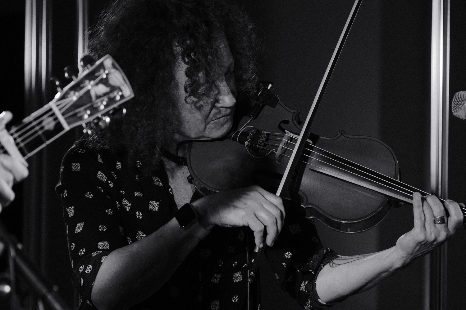 Black and white photo of a musician with curly hair playing violin, with a guitar visible in the background.