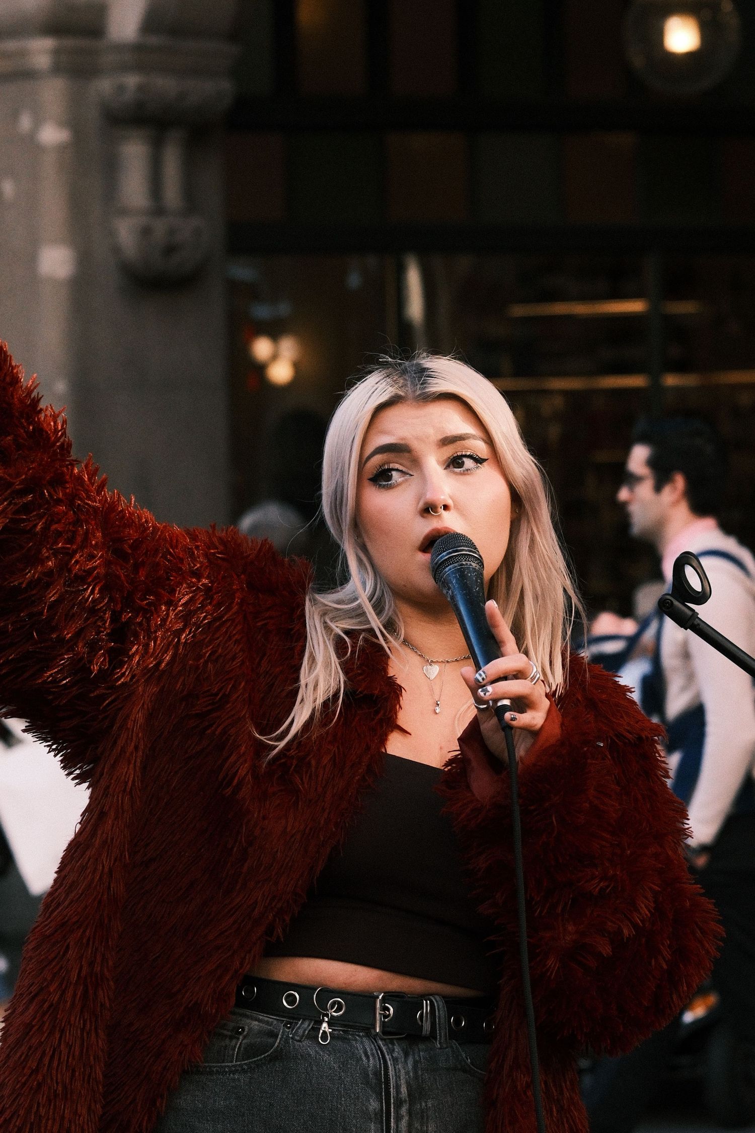 Young woman with blonde hair singing into microphone outdoors, wearing red fur coat and black crop top.