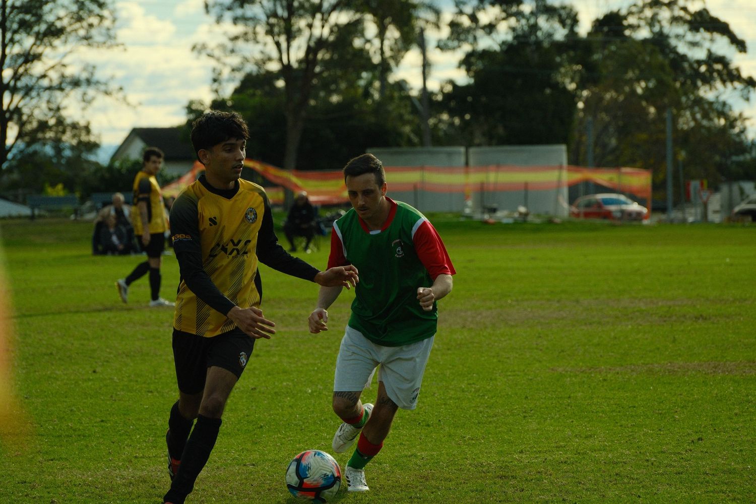 Two soccer players compete for the ball on a green field during an outdoor match, one in yellow and one in green jersey.