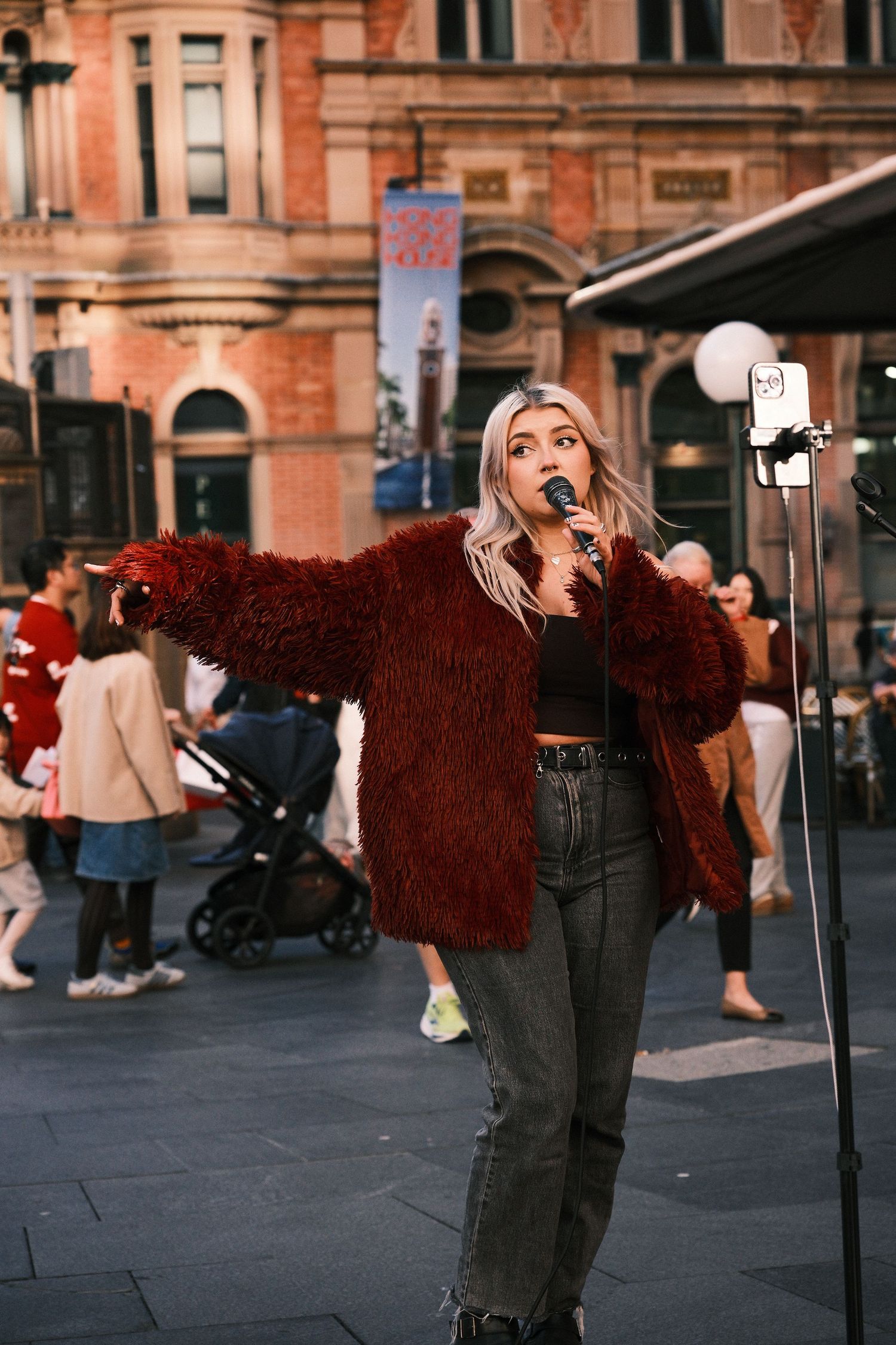 Blonde woman singing into microphone outdoors, wearing red fluffy jacket and crop top, performing on busy city street.