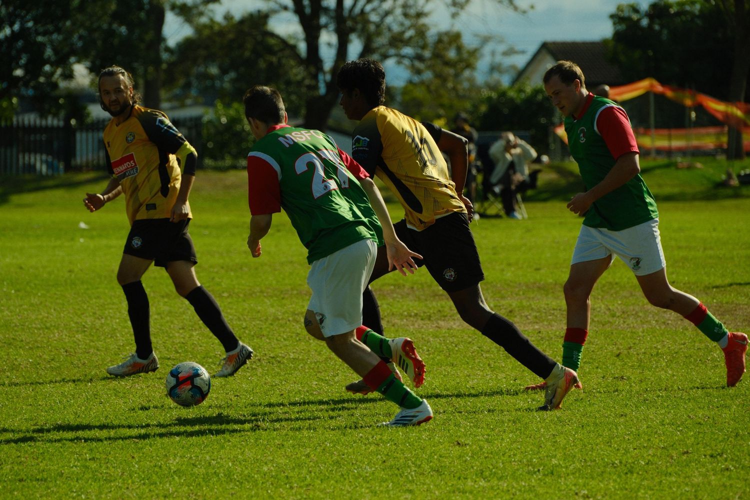 Soccer players in green and yellow jerseys compete for the ball on a sunny green grass field during a match.