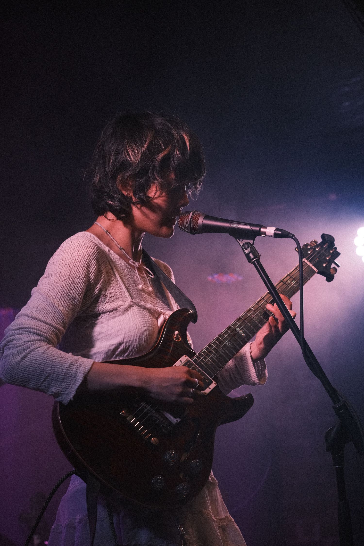 Female guitarist performing on stage with electric guitar at microphone under purple stage lighting.