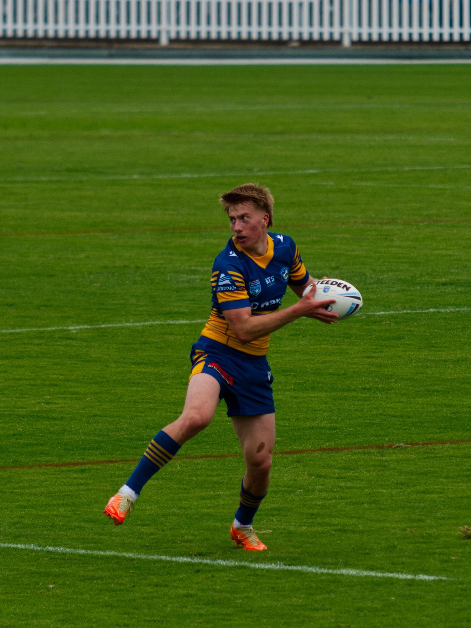 A young rugby league player in blue and gold jersey carries the ball on a field.