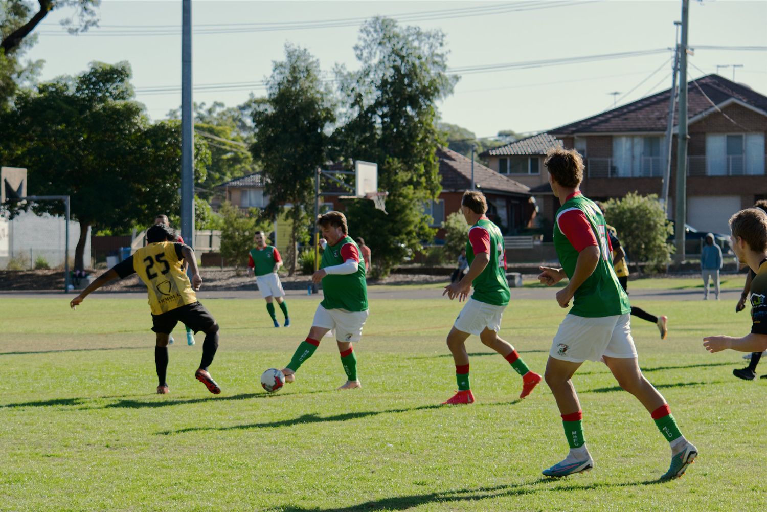 Soccer players in green and yellow uniforms compete on a sunny grass field during a match.
