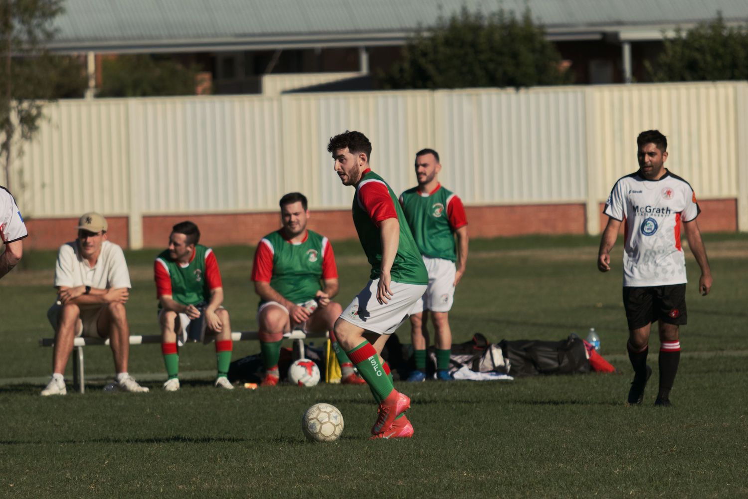 Soccer player prepares to kick the ball as teammates rest on the sideline during an outdoor match.