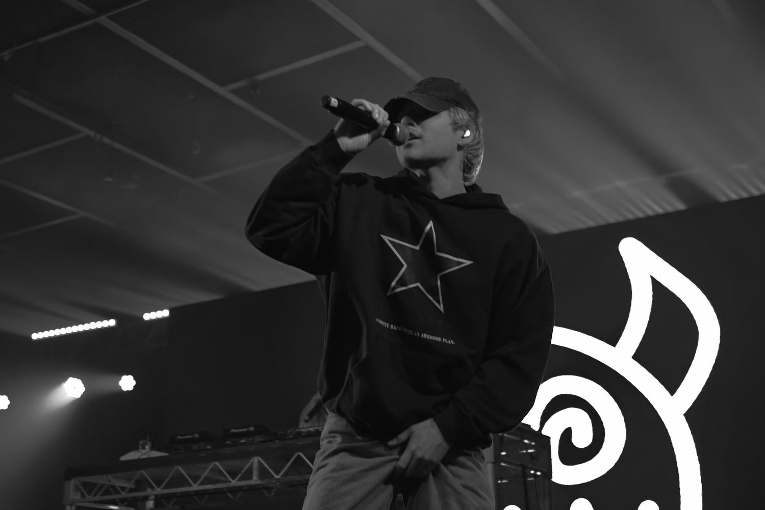 Male rapper sings into microphone wearing black star hoodie at live concert event.