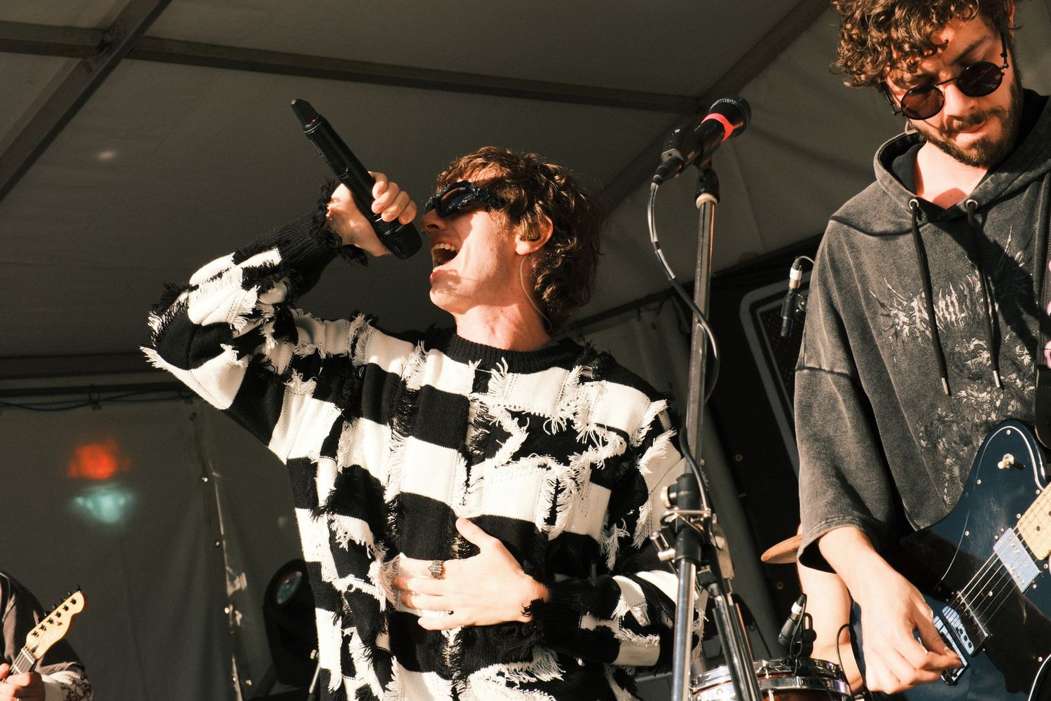 Singer in black and white patterned sweater performs on stage with band members playing guitars at outdoor concert.