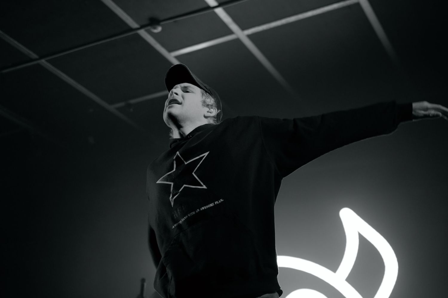 Person in black star sweatshirt poses dramatically under neon light in black and white photo.