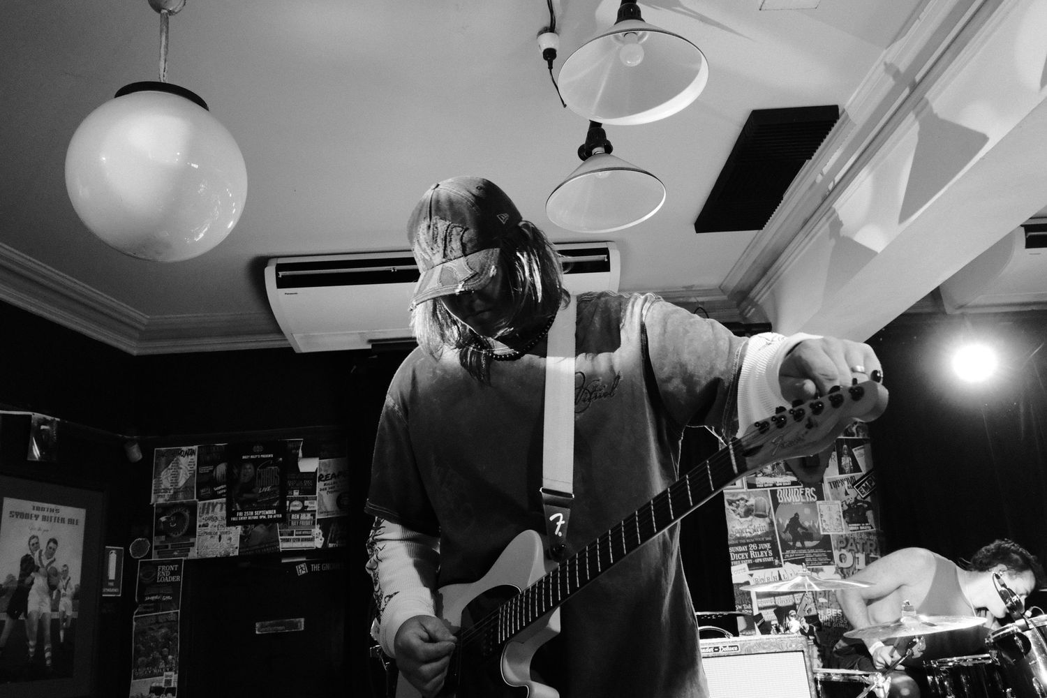 Black and white photo of a long-haired guitarist performing on electric guitar in a dimly lit indie music venue.