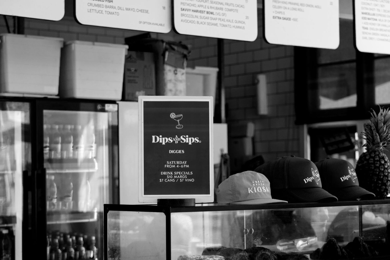 Black and white photo of Dips and Sips cafe counter with caps and a promotional sign on display.