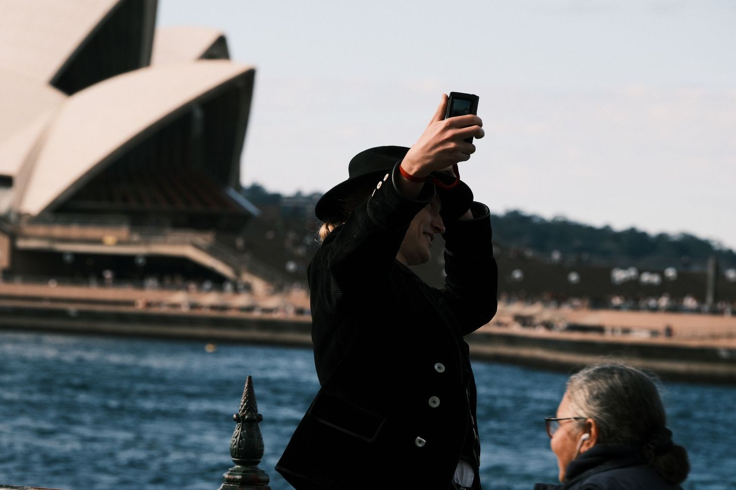 Tourist photographing Sydney Opera House with smartphone on a sunny day near the harbor waterfront.