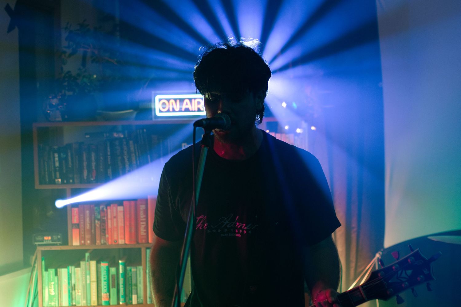 A musician performing on stage backlit by dramatic blue and purple beams with a bookshelf and ON AIR sign behind them.
