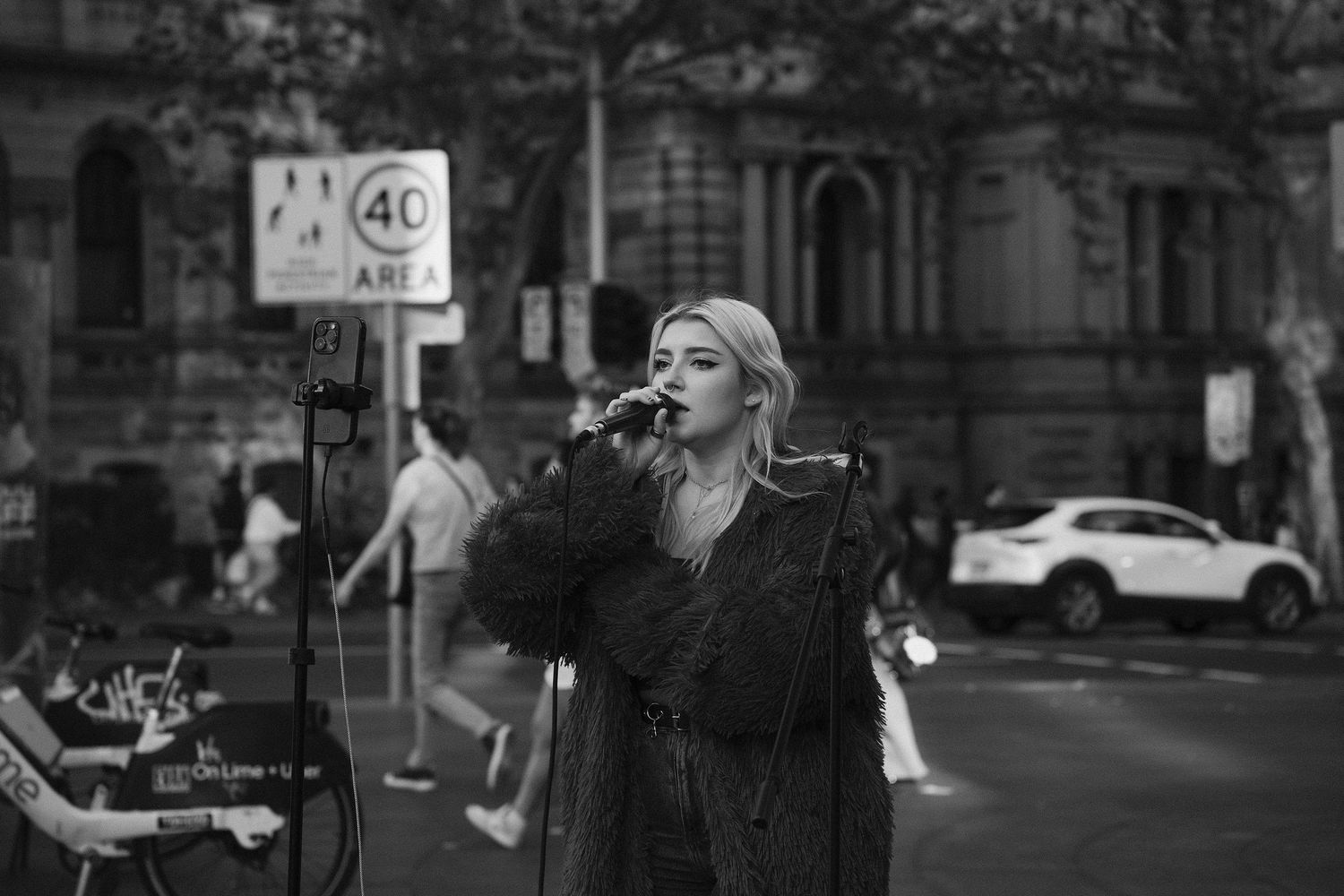 Black and white photo of a blonde woman singing into a microphone on a city street near a 40 speed limit sign.