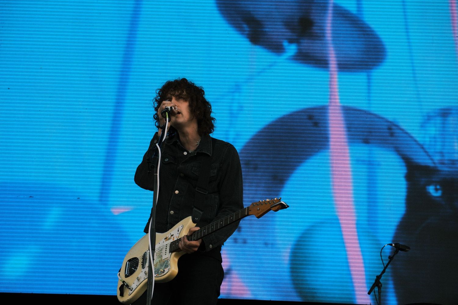 Male guitarist performing on stage with microphone, playing white electric guitar against blue backdrop at live concert.