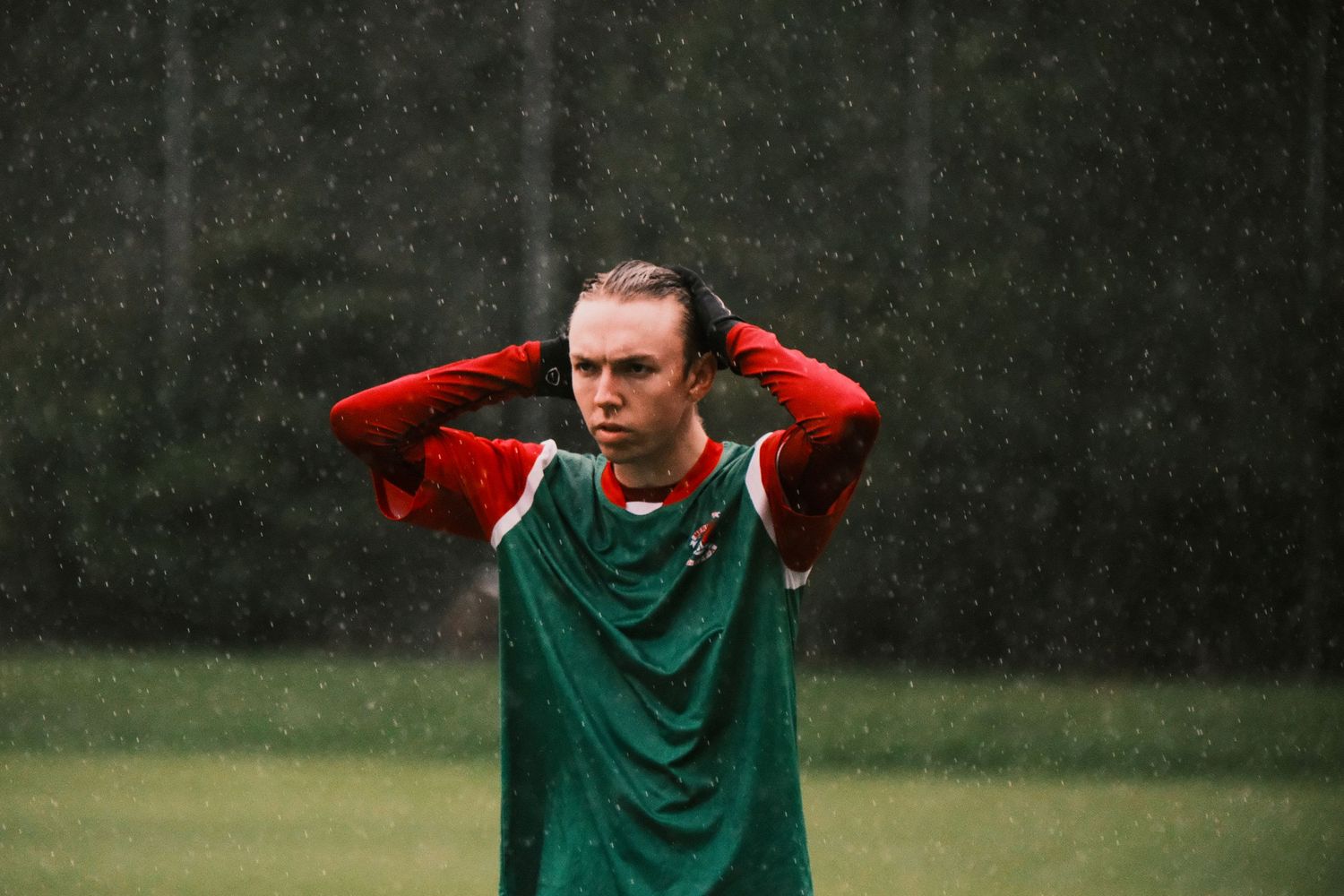 Soccer player in green and red kit stands in rain, hands behind head, on a training field.