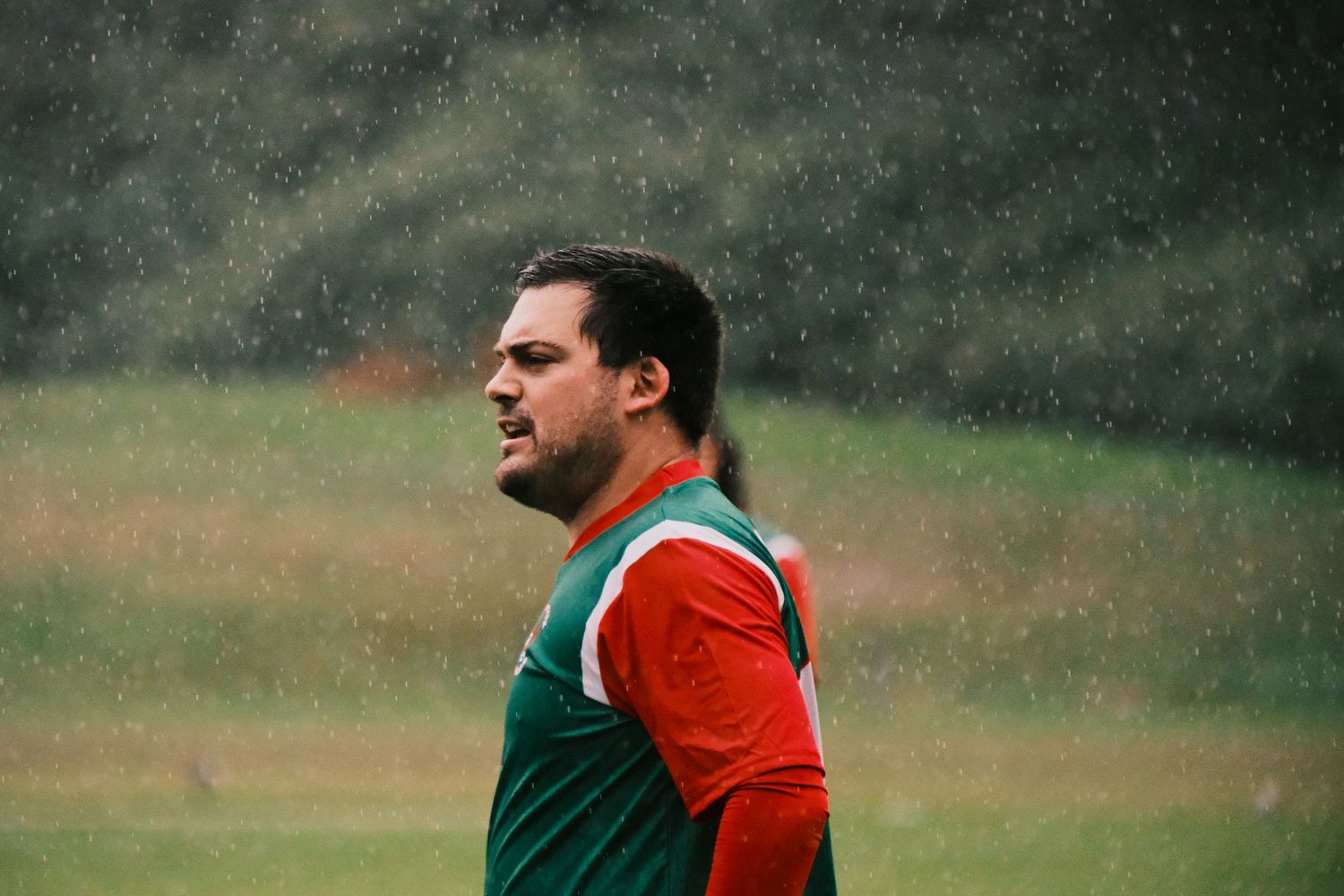 Man in red and green jersey standing outdoors in the rain during a sports training session.