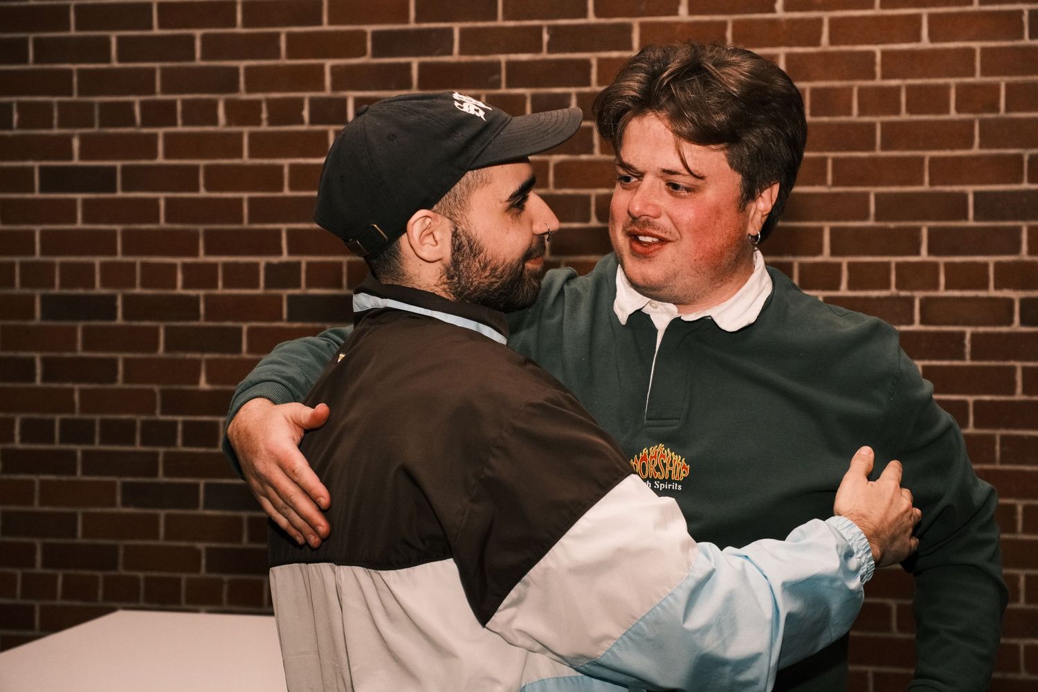 Two young men sharing a warm hug and smiling in front of a brick wall indoors.