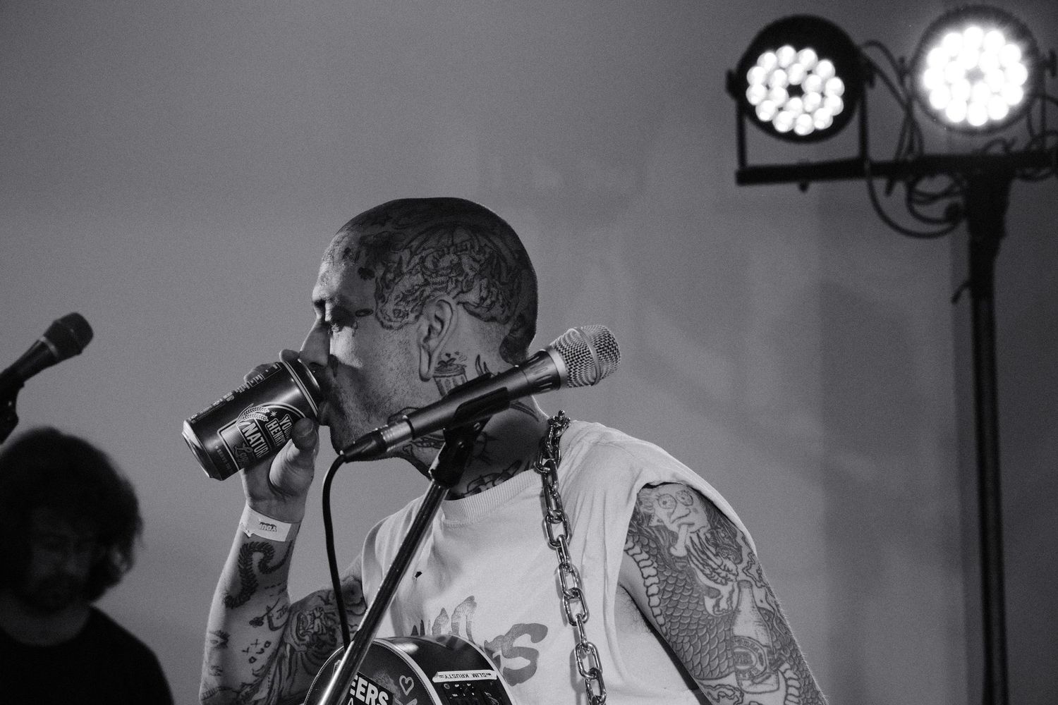 Tattooed male vocalist singing into microphone on stage in black and white concert photo with stage lights behind.