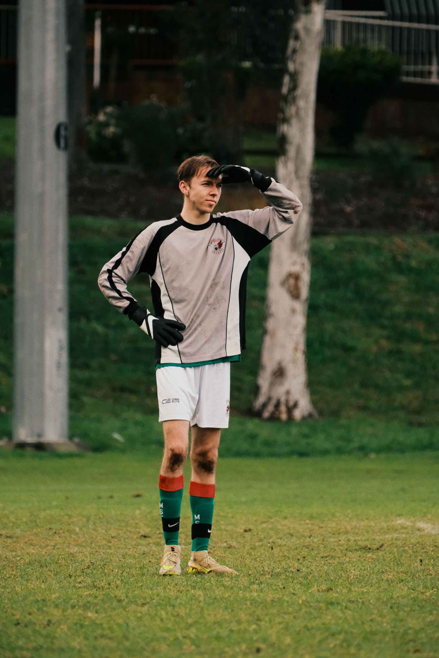 Young soccer goalkeeper in gray jersey, red and green socks, standing on grass field with gloves raised to forehead.