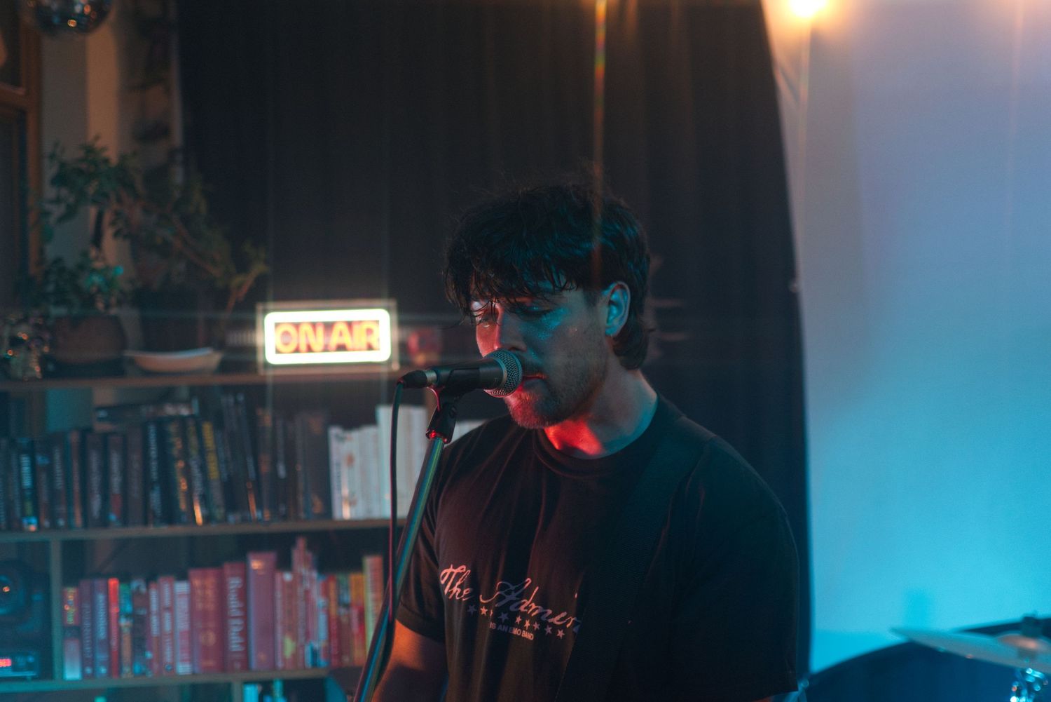 Young male vocalist performs at microphone in moody blue-lit room with bookshelf and neon sign.