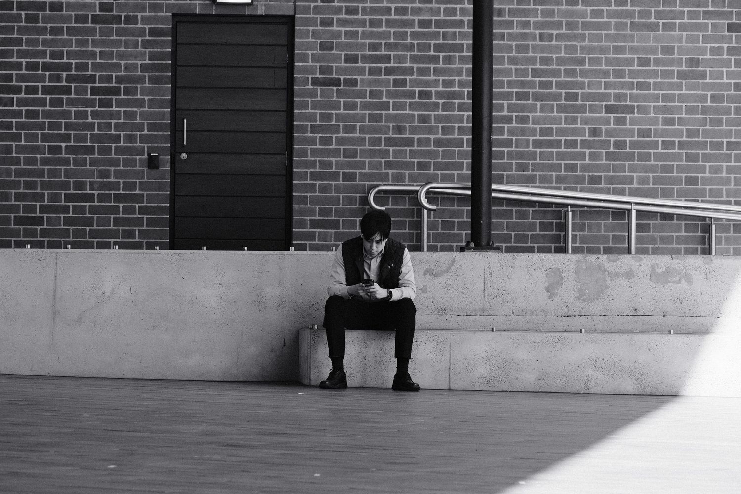 Black and white photo of a person sitting alone on a concrete bench outside a brick building, reading.
