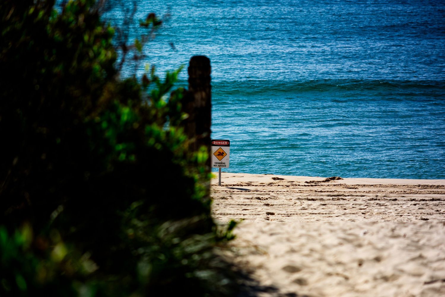 Sandy beach path leading to turquoise ocean waves with a warning sign and wooden post on a sunny day.