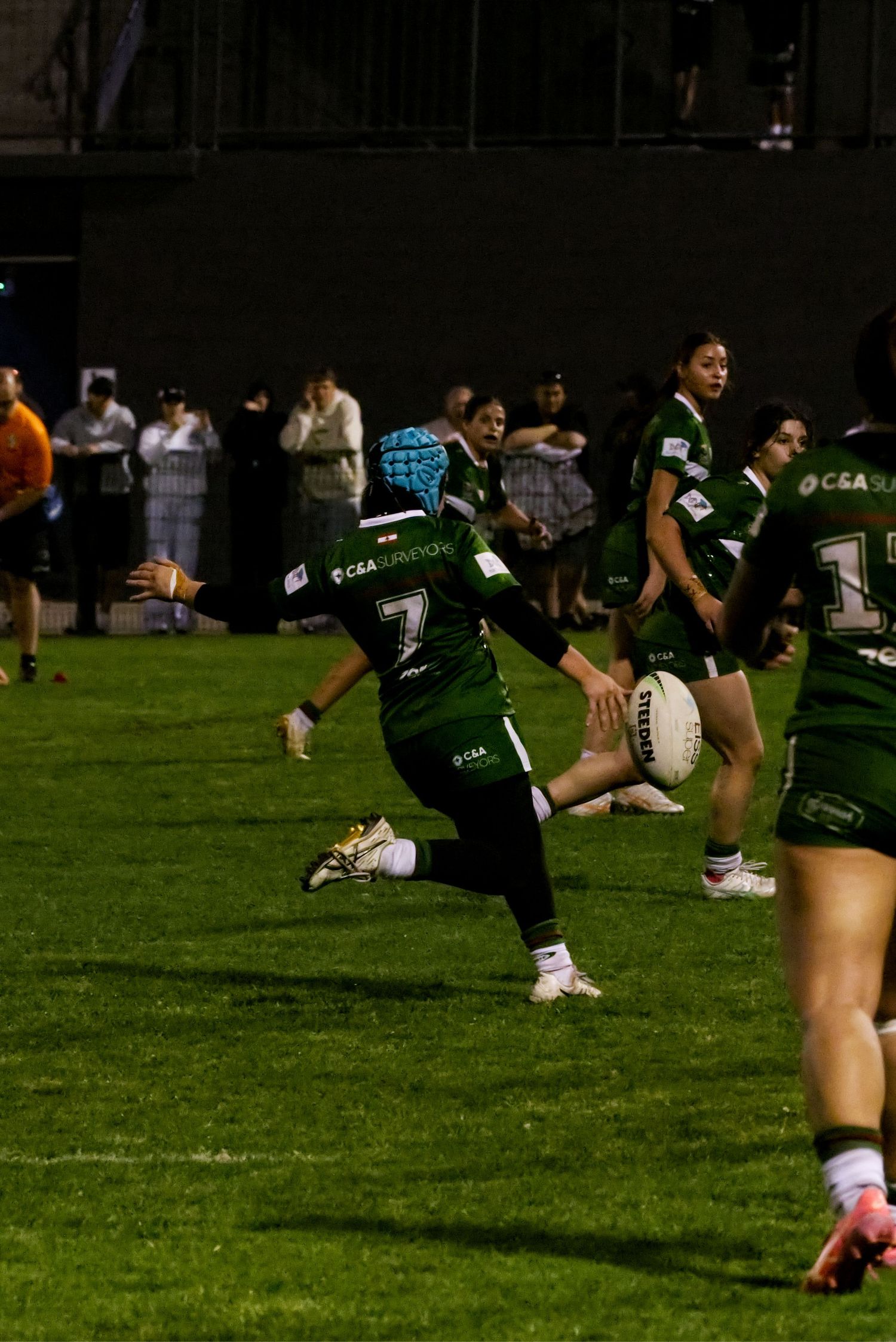 Women's rugby league player number 7 in green kit kicks ball during night game with crowd watching.