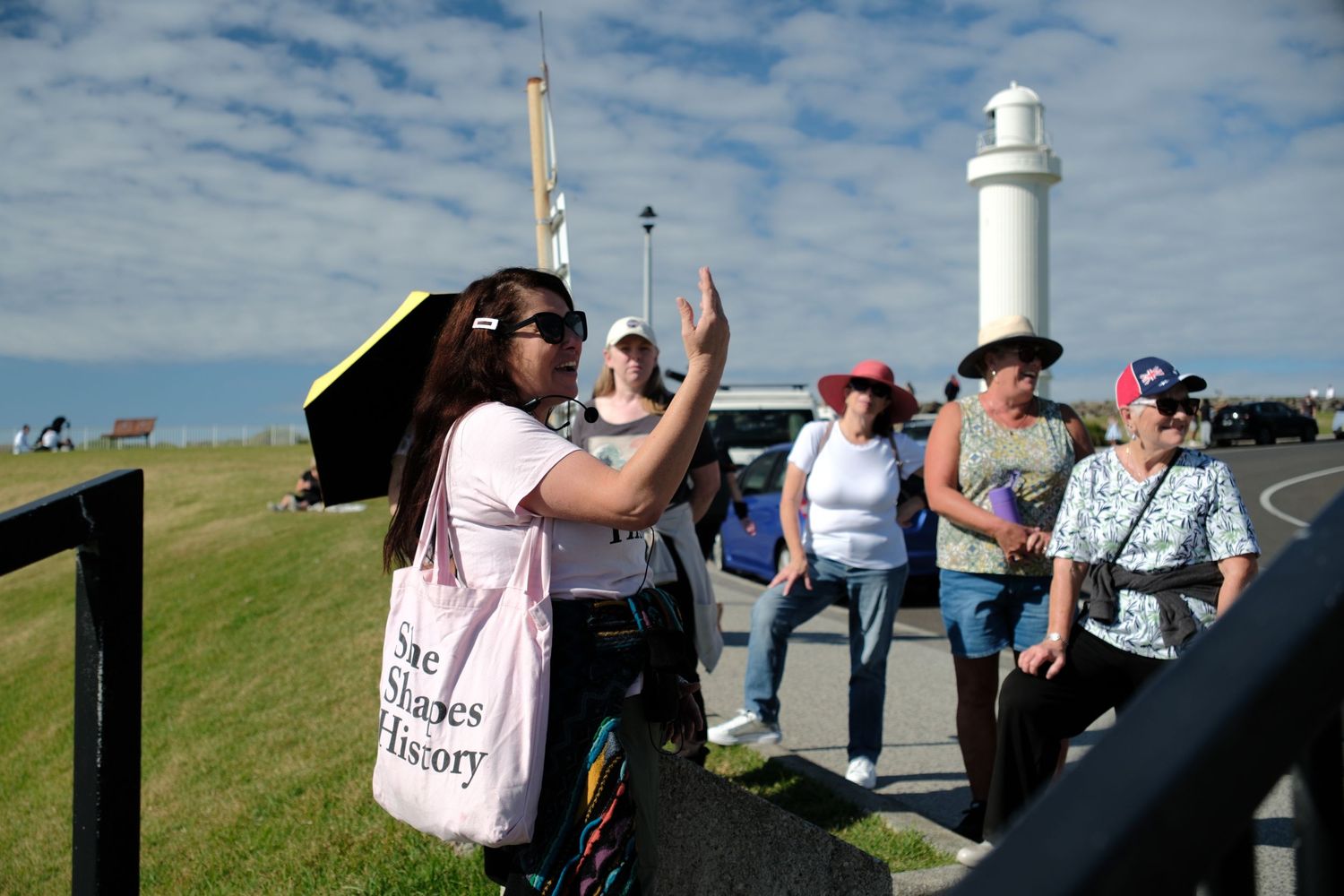 Tour guide gesturing to a group of visitors near a white lighthouse on a sunny day.