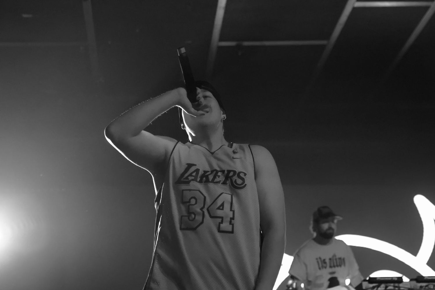 Hip-hop artist in Lakers 34 jersey performs on stage with microphone at live show.