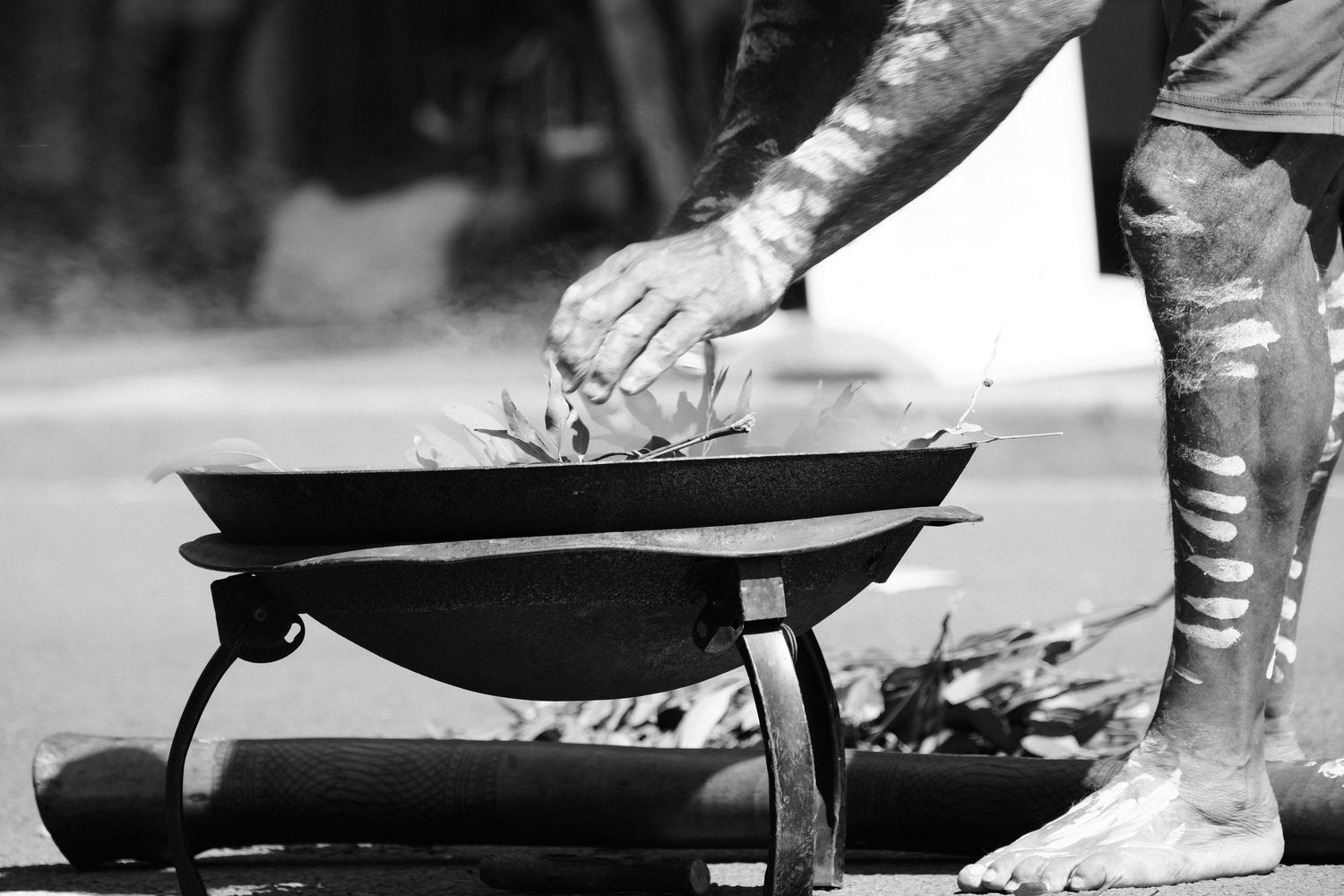 Aboriginal man with painted body tending a traditional smoking ceremony fire in a black and white photo.