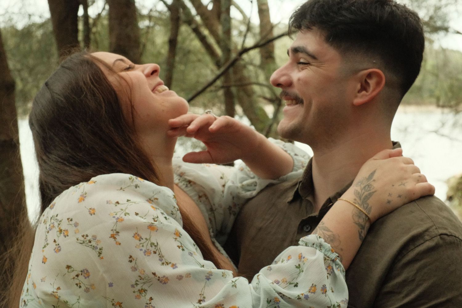 A joyful couple laughs and embraces outdoors among trees, the woman in a floral dress and man in an olive shirt.