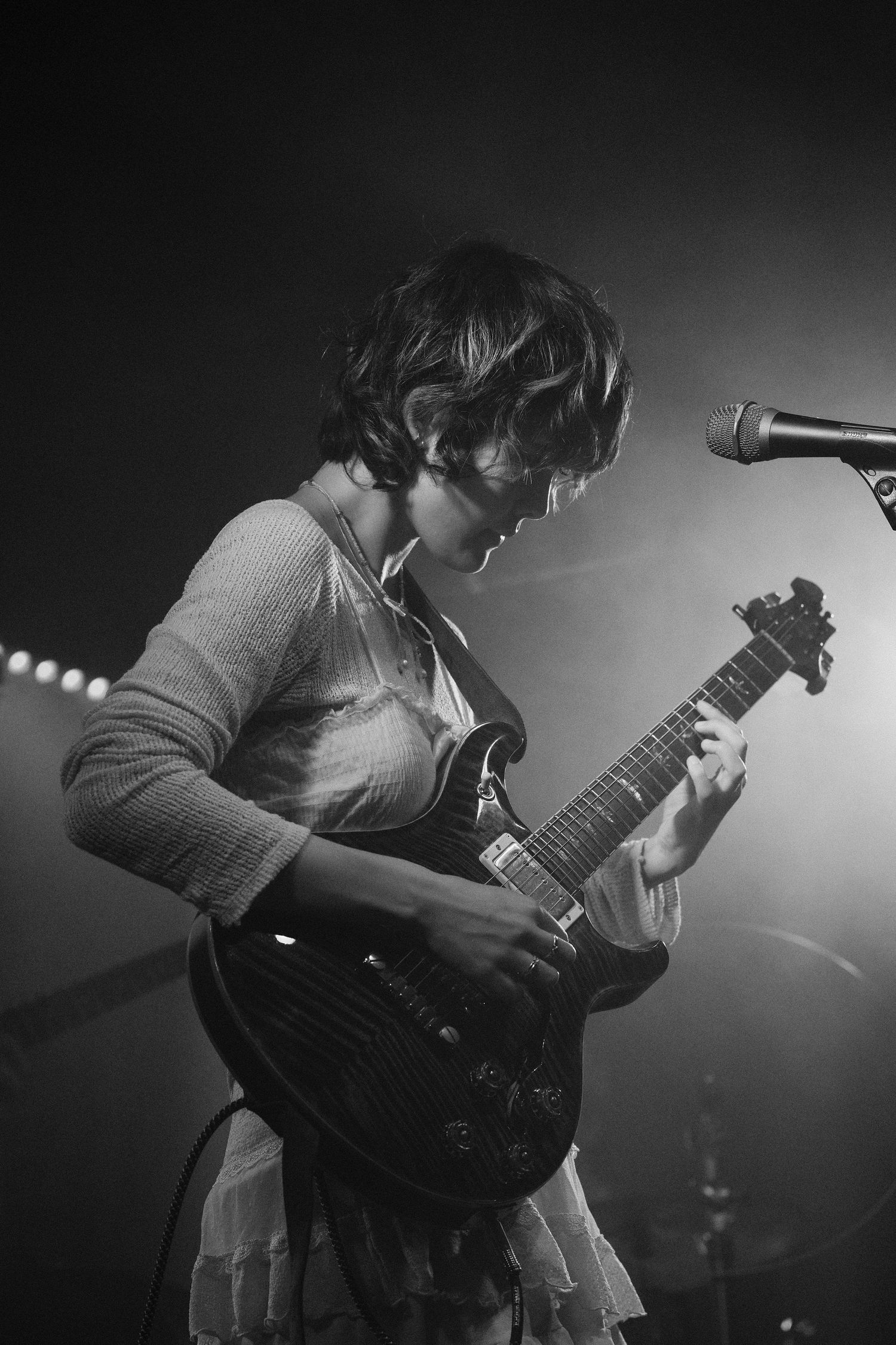 Black and white photo of a young woman playing electric guitar on stage under dramatic lighting.