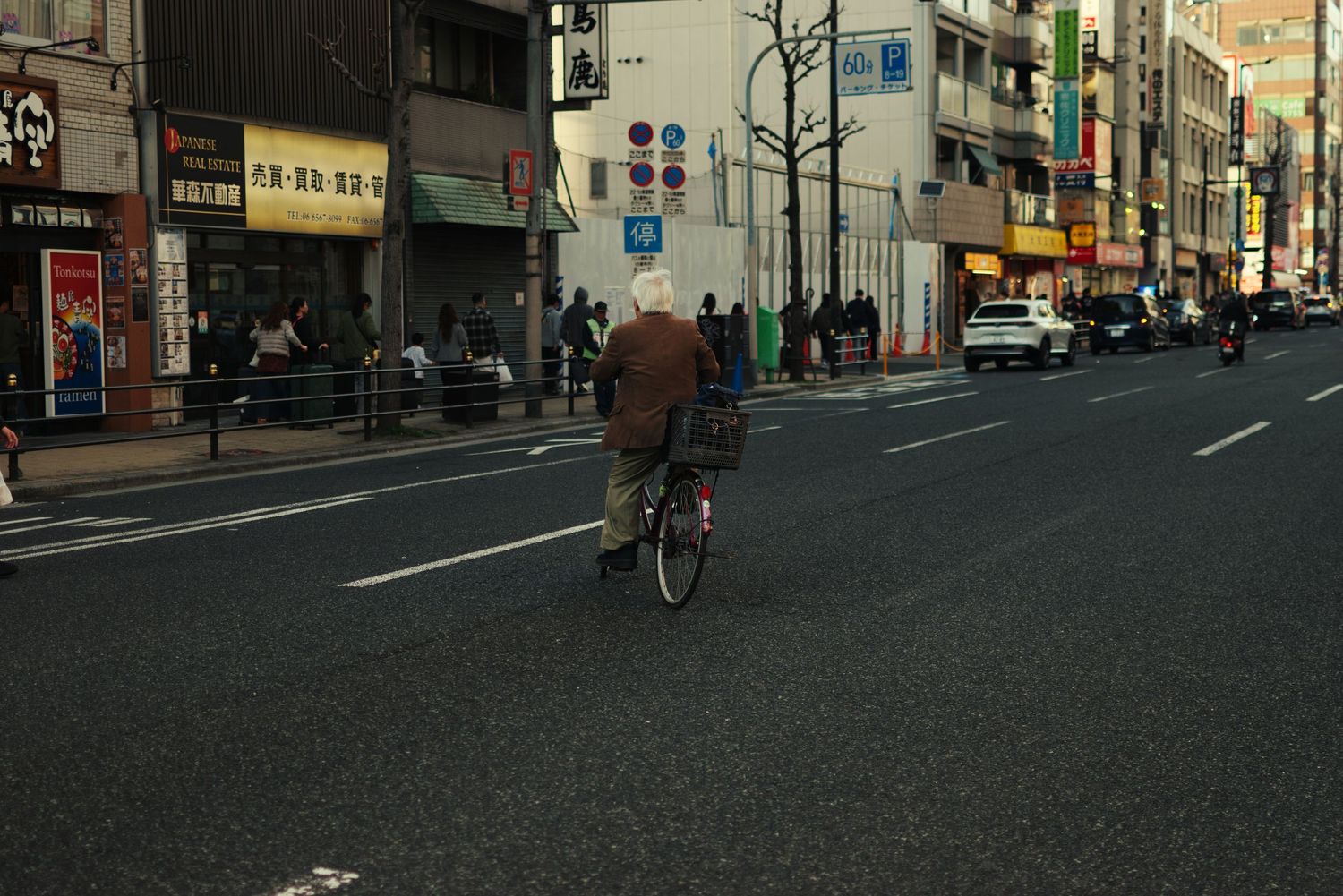 An elderly person rides a bicycle across a wide urban Japanese street at dusk.