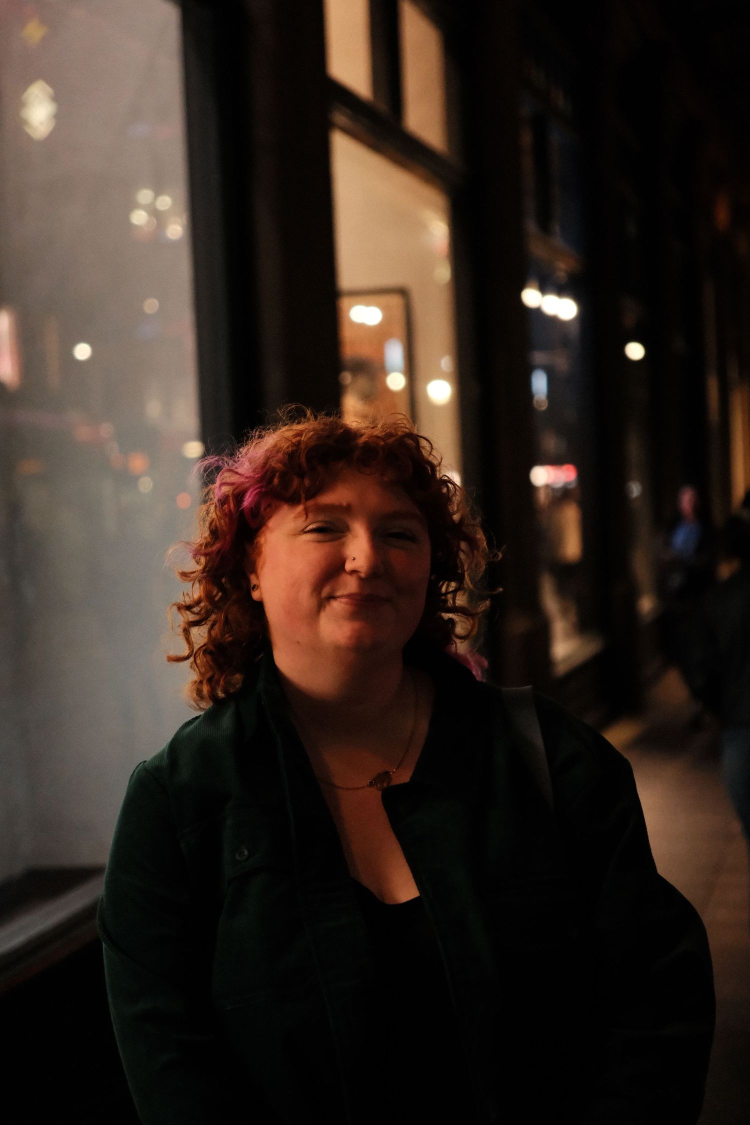 Woman with curly red hair smiling at night outside a lit storefront.