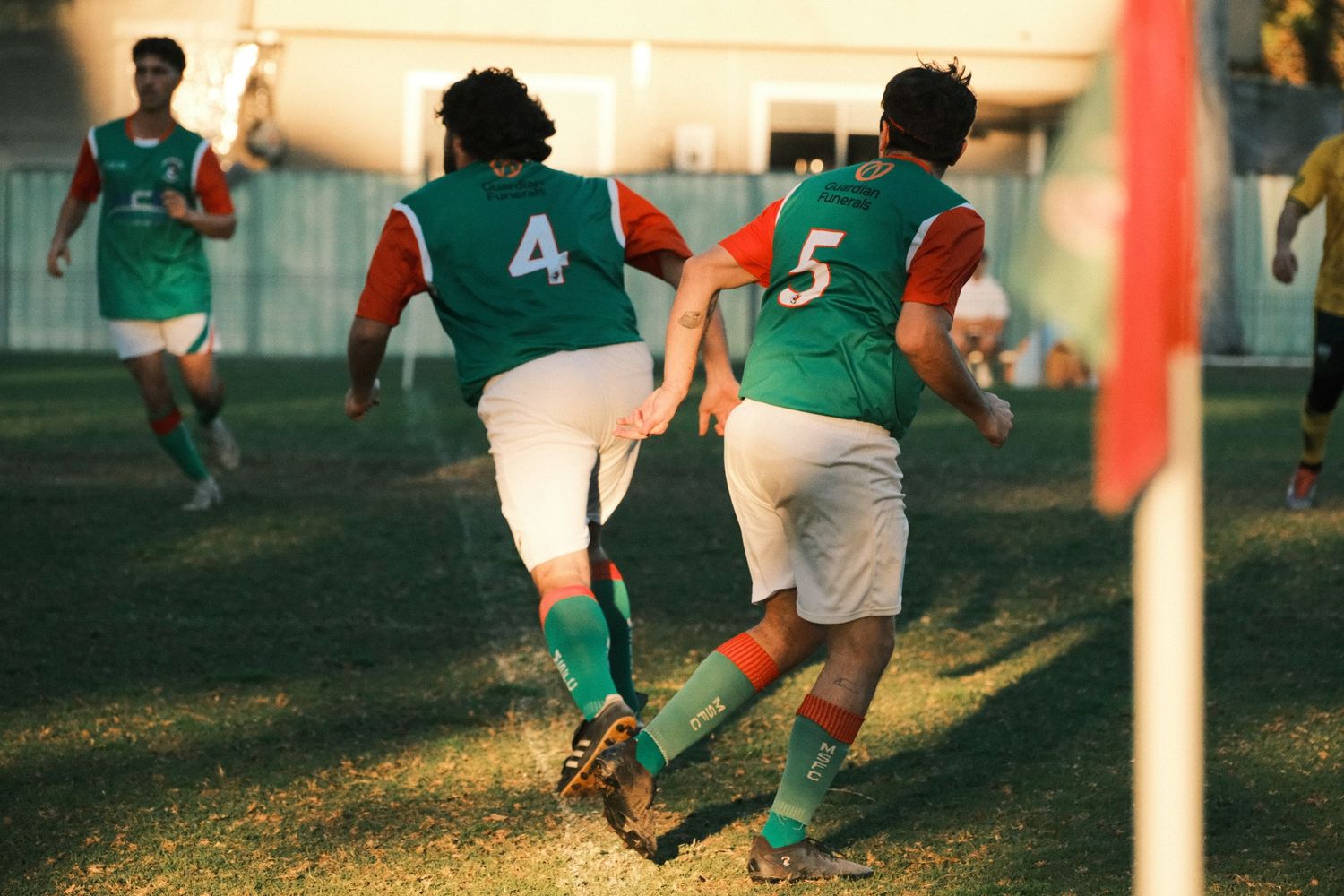 Soccer players in green and orange jerseys numbered 4 and 5 compete near a corner flag on a grass field.