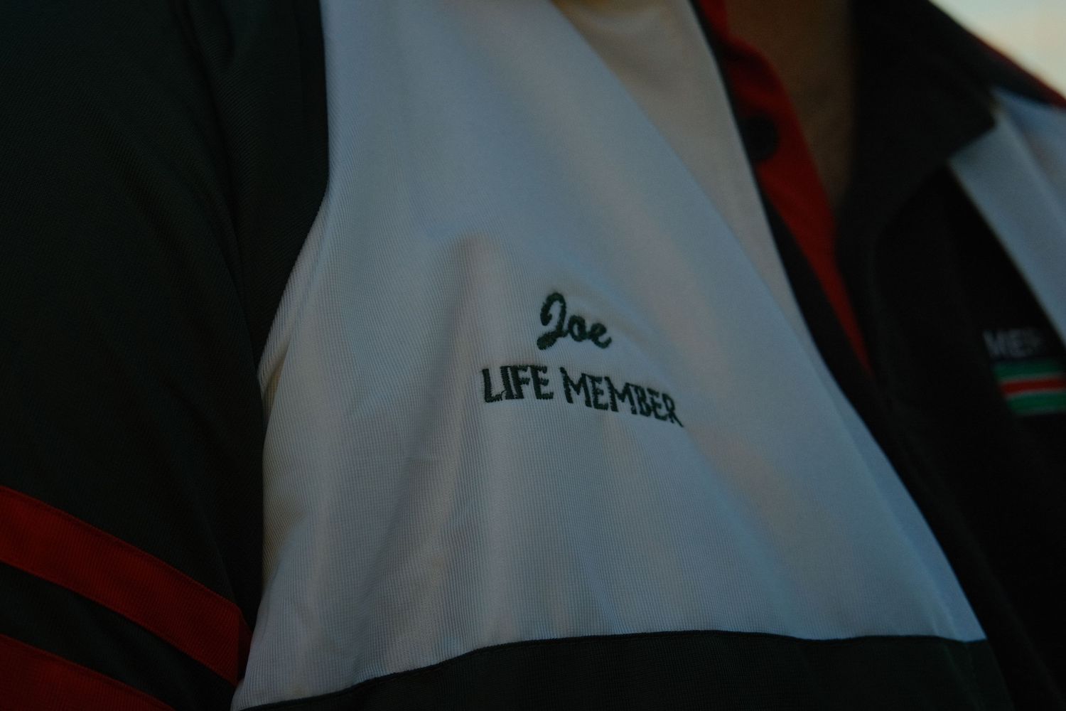 Close-up of a white polo shirt embroidered with Joe Life Member in green text.