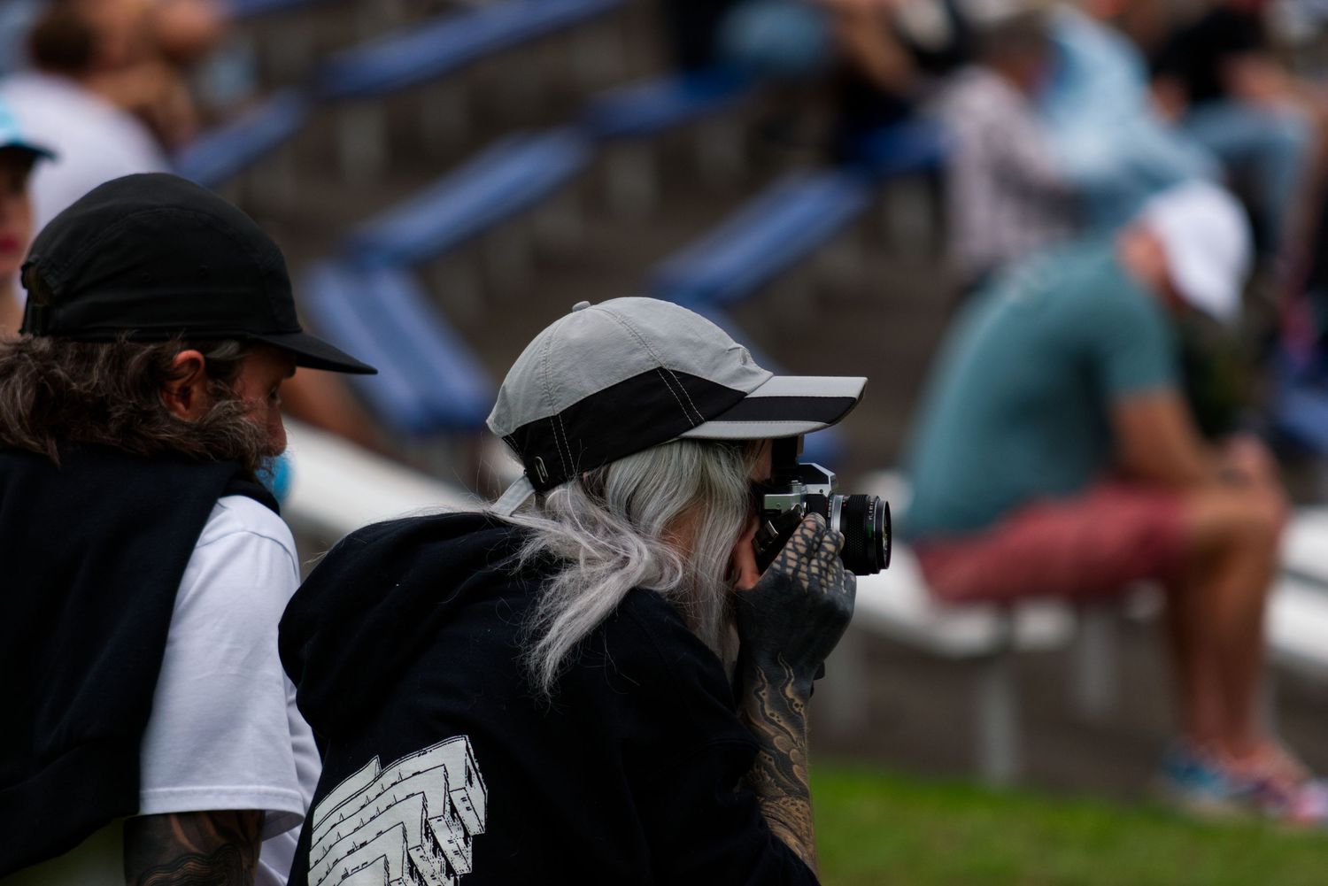Woman with white hair wearing a cap uses camera at an outdoor stadium.