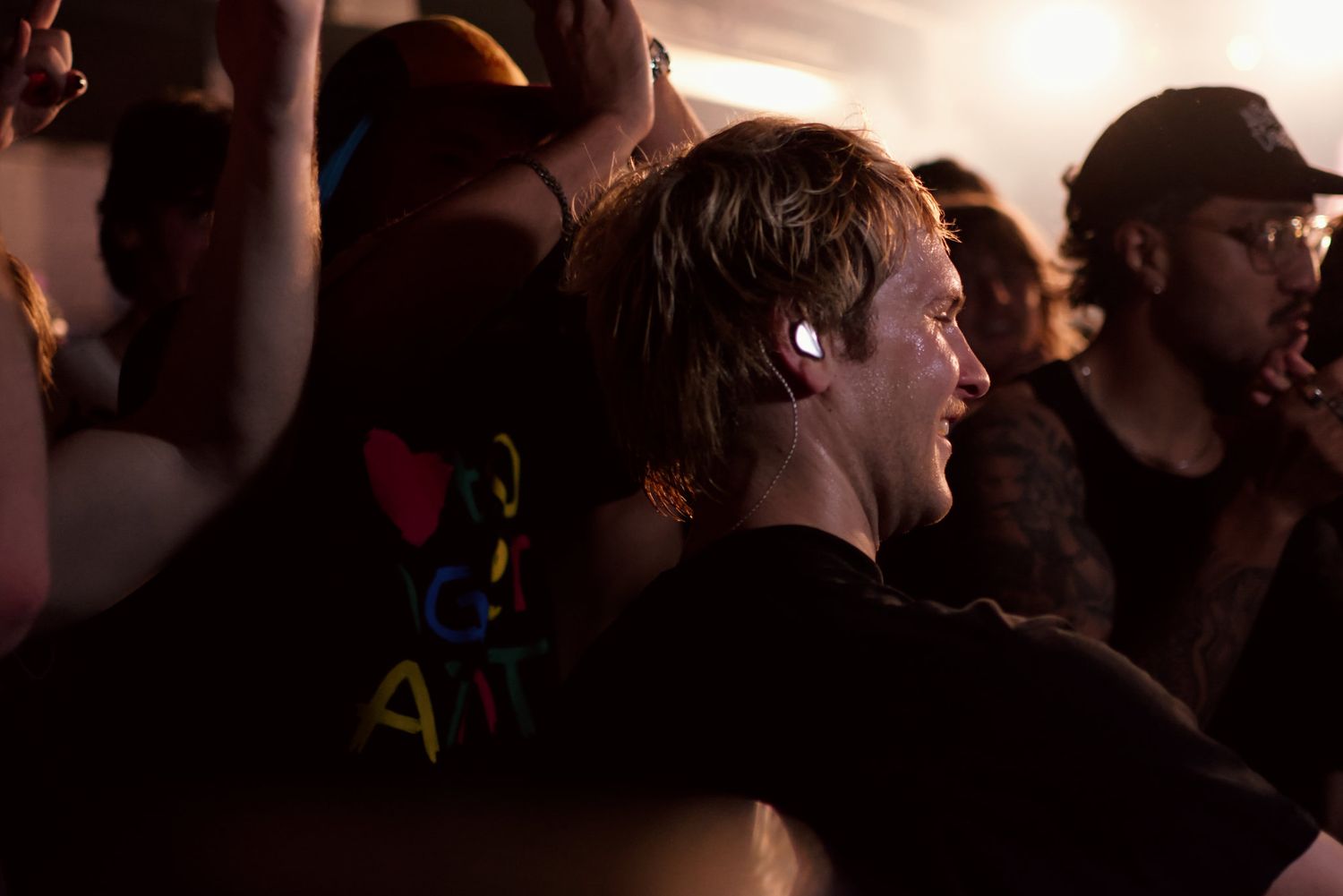 Concert attendee smiles in crowd wearing black tee with glowing earbuds at live show.