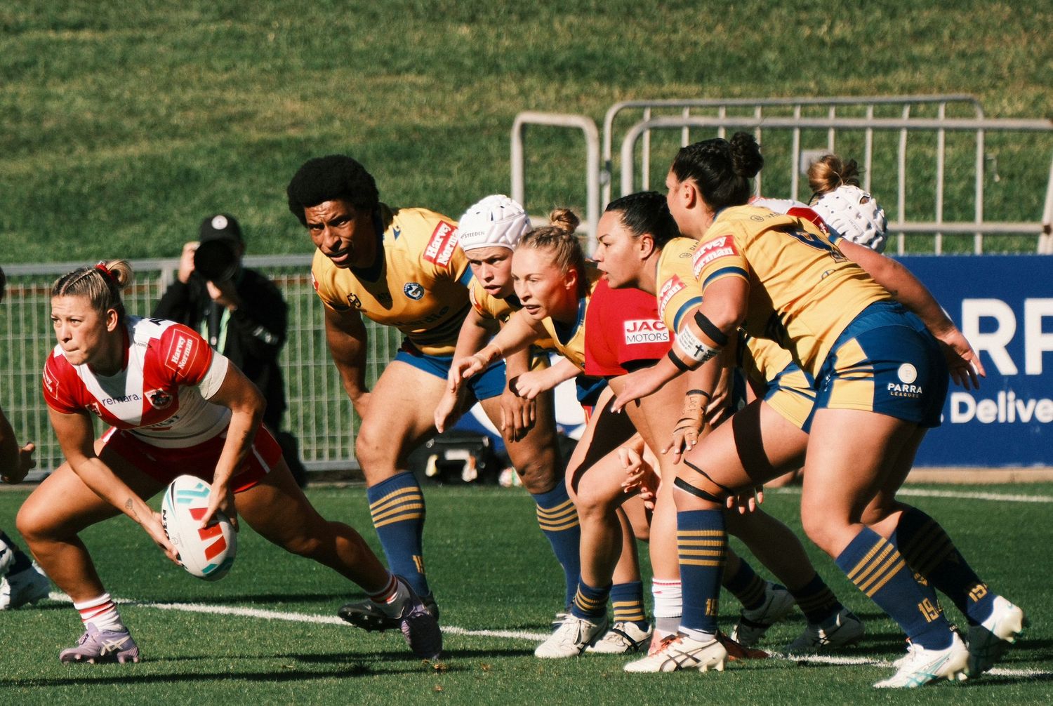 Women rugby players compete in an intense match, with players in yellow and red uniforms scrummaging for the ball on a green field.