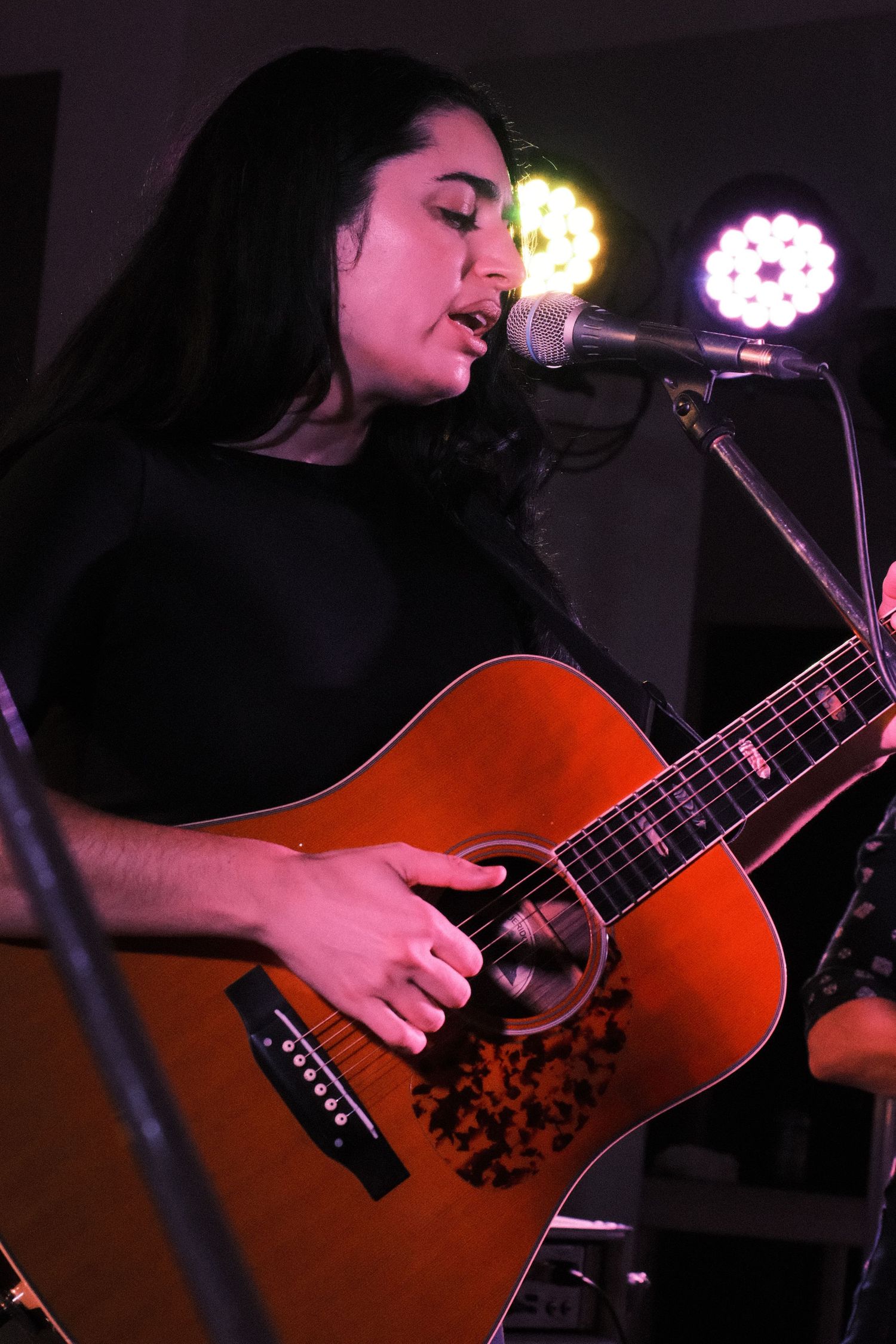 A young woman with long dark hair plays an acoustic guitar and sings into a microphone at a live music performance.