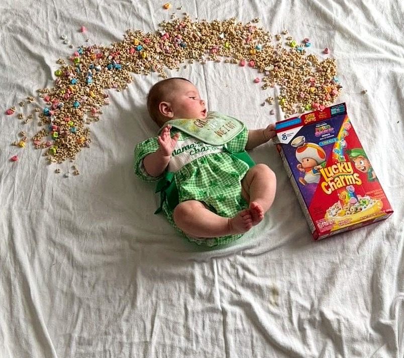 Sleeping baby in green outfit beside Lucky Charms box with cereal arc.